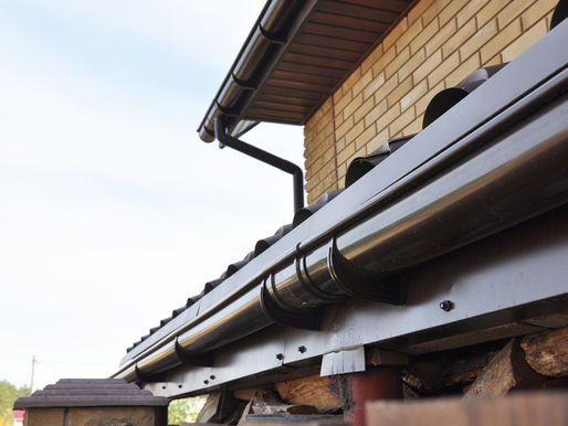 Close-up of a house roof gutter and downspout against a brick wall and pale sky.