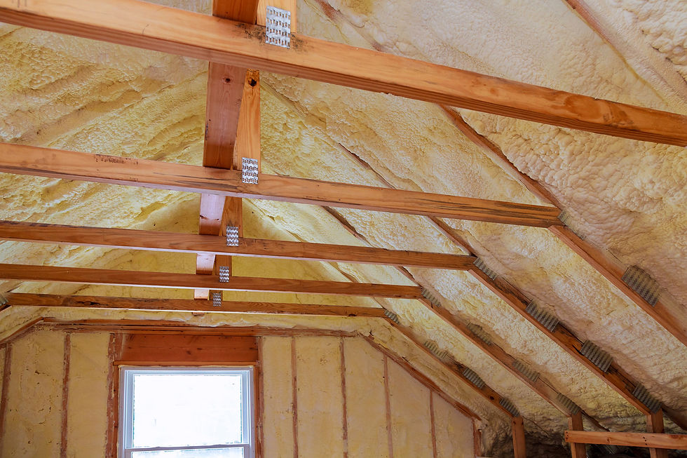 Attic interior with exposed wooden rafters, yellow spray foam insulation, and a small window below, showing unfinished construction.
