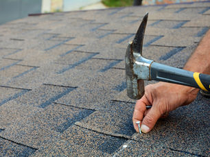 Hand hammering a nail into brown roof shingles. Close-up of textured surface. No text. Outdoor daytime setting.