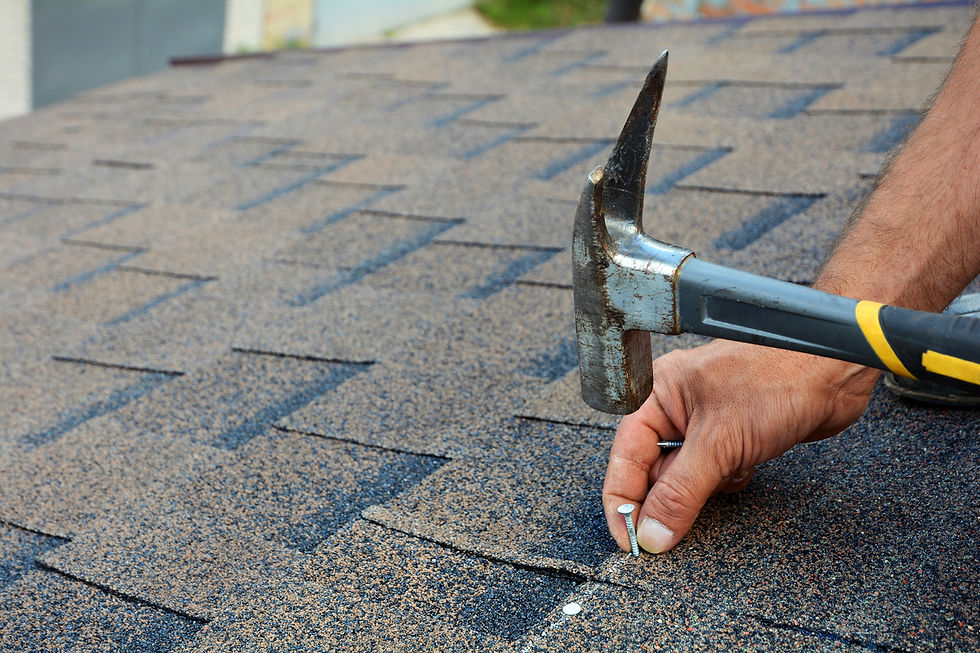 Hand using a hammer to nail gray shingles on a roof. Close-up shows textured pattern. Bright outdoor setting, focused on detailed work.