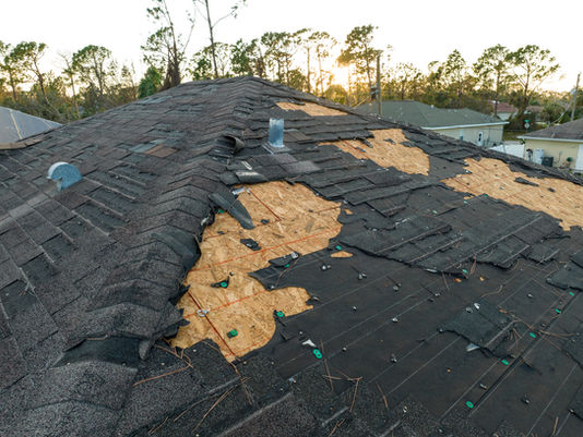Damaged roof with missing shingles and exposed wooden areas against a backdrop of trees and a sunset.