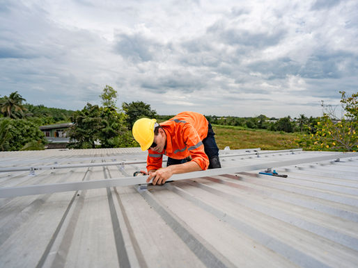 Worker in orange safety gear and yellow hard hat installs solar panels on a metal roof under a cloudy sky, with green fields behind.