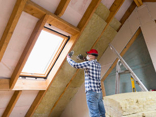 Man in red cap and plaid shirt installing insulation in an unfinished attic with skylight, ladder, and exposed wood beams.