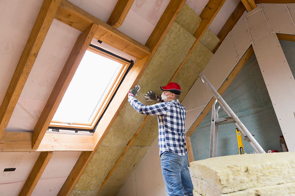 Man in red cap and plaid shirt installing insulation in an unfinished attic with skylight, ladder, and exposed wood beams.