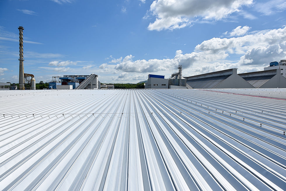 Wide view of a corrugated metal factory roof with smokestack and industrial buildings under a bright blue, cloudy sky