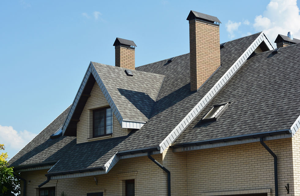 Brick house with grey shingled roof, skylights, and chimneys against a blue sky. Bright, sunny day with a few clouds.