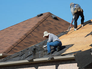 Two workers repair shingles on a sloped roof. One kneels, while the other uses a tool. Clear sky and red-brown shingles are visible.