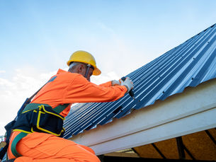 Worker in an orange suit and yellow helmet secures a blue metal roof with a drill. Clear sky in the background.
