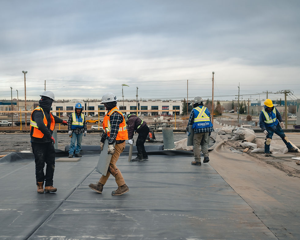 Construction workers in hard hats and safety vests install roofing membrane on a flat roof under a cloudy sky, with one vest reading SUPERIOR