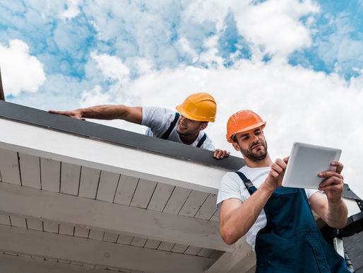 Two construction workers wearing orange and yellow helmets on a rooftop. One uses a tablet, blue sky with clouds in the background.
