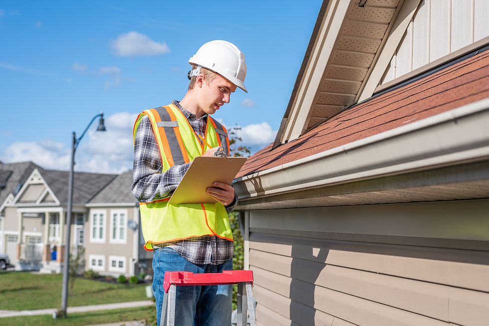 A construction worker in a hard hat and safety vest inspects a roof with a clipboard. He's on a ladder outside suburban houses under a blue sky.