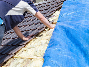Worker installing brown roof tiles over insulation beside a blue tarp on a sunny day.
