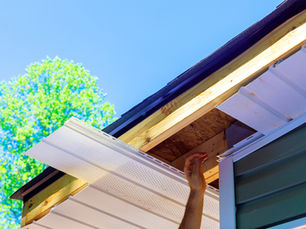 Hand installing white soffit panels under a house roof eave, with blue sky and green tree in the background.