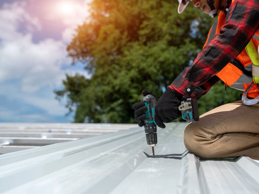 Worker in a red plaid shirt and orange vest using a drill on a best roofing material, metal roof, under a sunny sky with trees in the background.