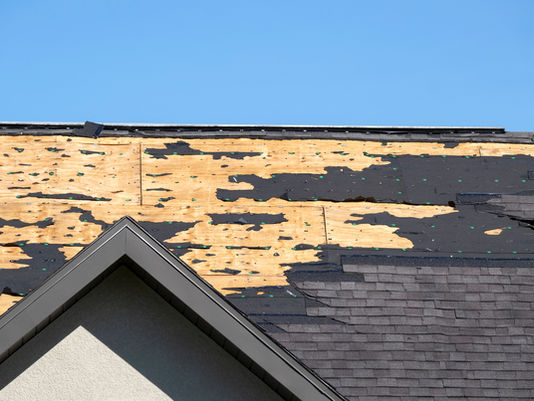 Damaged roof with peeling shingles and exposed wood above a house gable, set against a clear blue sky.