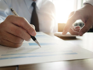 Close-up of a businessman writing on charts while another hand points at a document in a bright office.