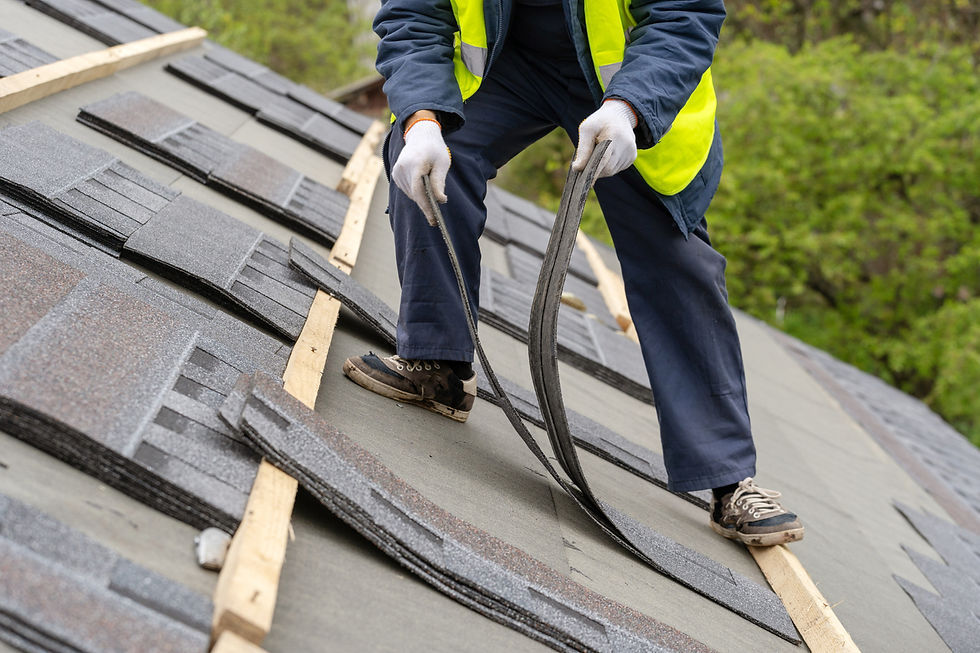 A roofing worker in a safety vest and gloves installs shingles on a sloped roof, with trees in the background.
