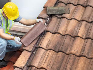 Worker in yellow hard hat and vest repairing brown roof tiles on a sloped roof. Sunny setting, focus on intricate tile placement