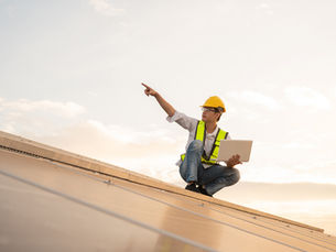 Construction worker on a roof in a yellow hard hat and reflective vest holds a tablet, pointing into the distance, under a bright sky.