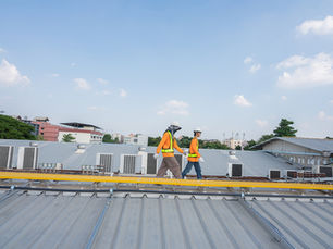 Two workers in orange vests and helmets walk on a metal roof. Clear sky and buildings in the background. Safety gear visible.