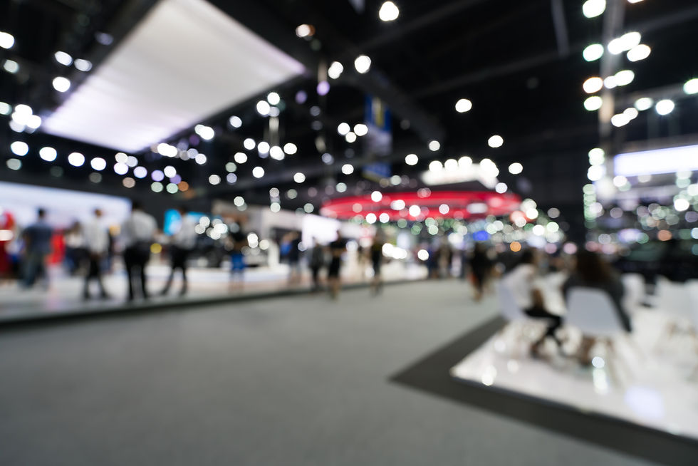 Blurry trade show hall with people browsing booths under bright overhead lights, creating a busy, modern expo atmosphere.