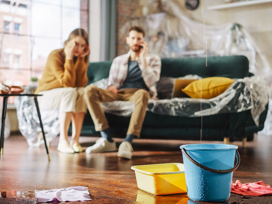 Couple sits on a couch in a water-damaged living room, with buckets, towels, and plastic sheeting on the floor.