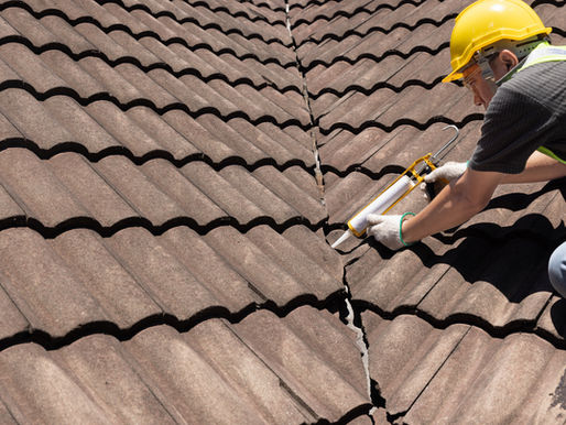 A worker in a yellow helmet and gloves repairs a cracked brown tile roof with sealant. Sunny day.
