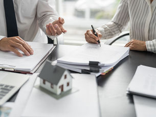 Two professionals review and sign real estate documents at a desk, with a house model, laptop, and paperwork in a bright office.