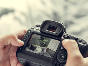 Hands holding a camera, screen showing a bedroom with plants and brick wall. Soft lighting, buttons visible, creating a calm mood for professional photography.