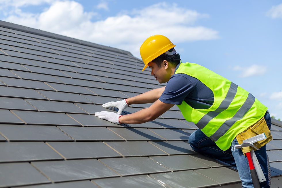 Worker in yellow hard hat and vest repairing a roof under a blue sky. He uses gloved hands to adjust tiles, with tools on his belt.