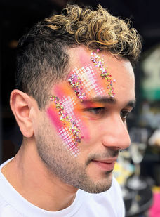 Man with orange pnk and white abstract painted design with gold glitter on the side of his face