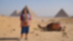 Bettany Hughes stands in a desert landscape; a camel rests nearby, with the pyramids rising in the distance.