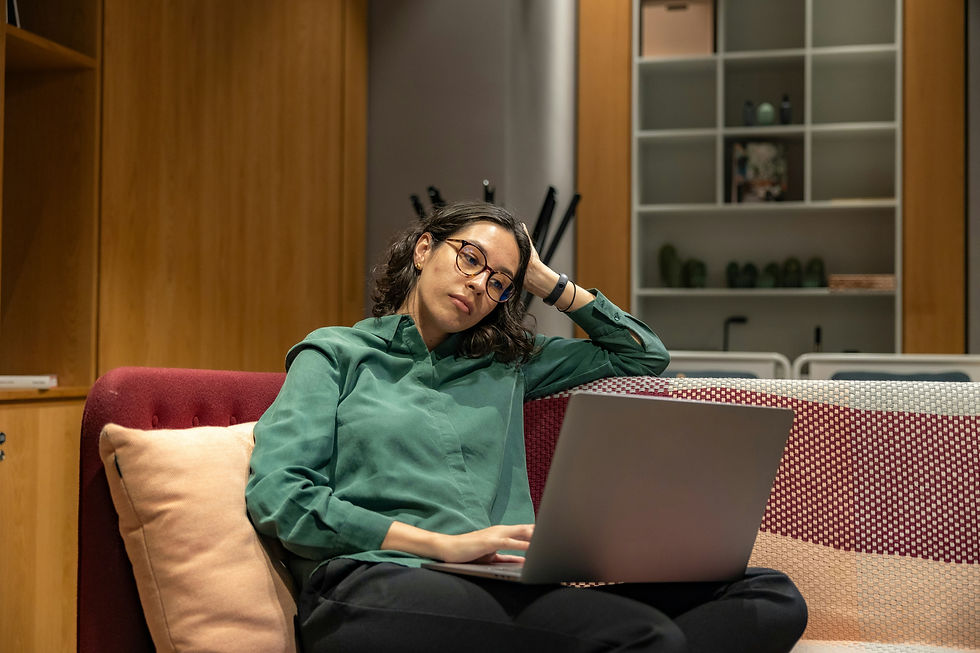 Woman in glasses sitting on a couch with a laptop, resting her head on her hand and looking fatigued or disengaged, suggesting burnout or mental exhaustion while working.