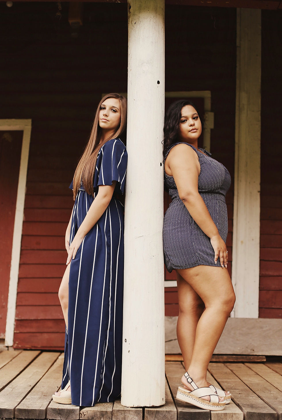 Two women standing back-to-back against a white pillar on a rustic porch, each with a calm, confident expression—celebrating body diversity, individuality, and strength in stillness.