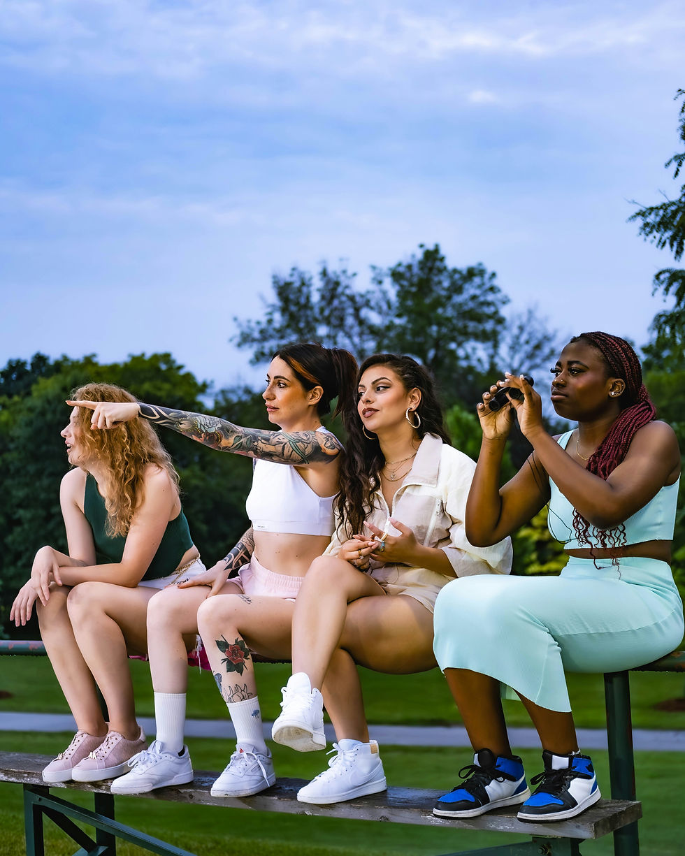 Group of four diverse women sitting on bleachers outdoors, casually dressed, one pointing into the distance while another uses binoculars—capturing connection, curiosity, and shared focus in a relaxed social setting.