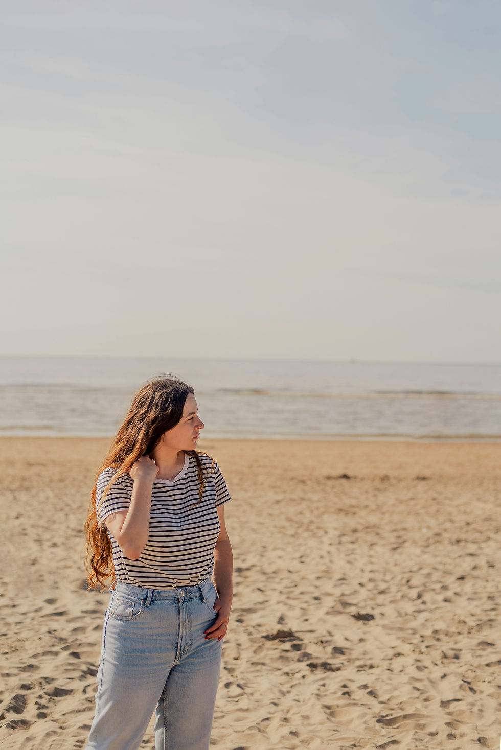 A woman standing on a quiet beach, gently holding her hair and gazing into the distance—capturing a moment of reflection, calm, and inner stillness.