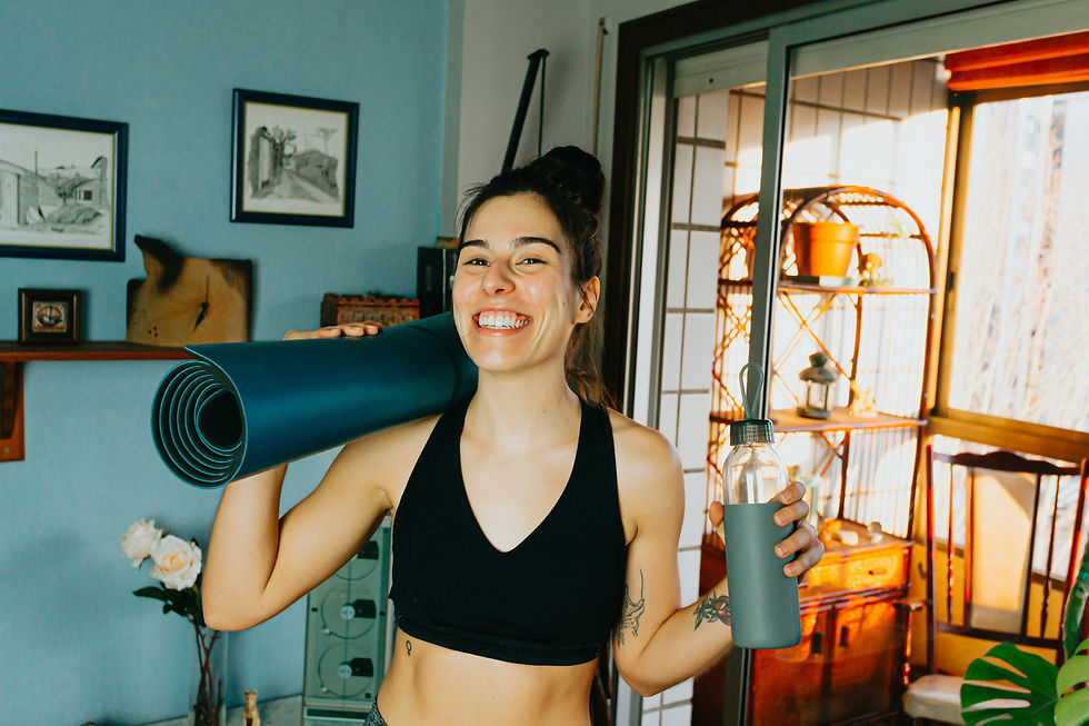 A smiling woman in activewear stands indoors holding a rolled-up yoga mat and a water bottle, radiating energy and readiness for movement or a workout.