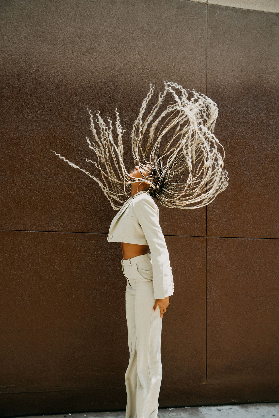 A confident woman in a cream suit flipping her long braids back in mid-air, standing tall against a plain brown wall—capturing freedom, self-expression, and embodied strength.