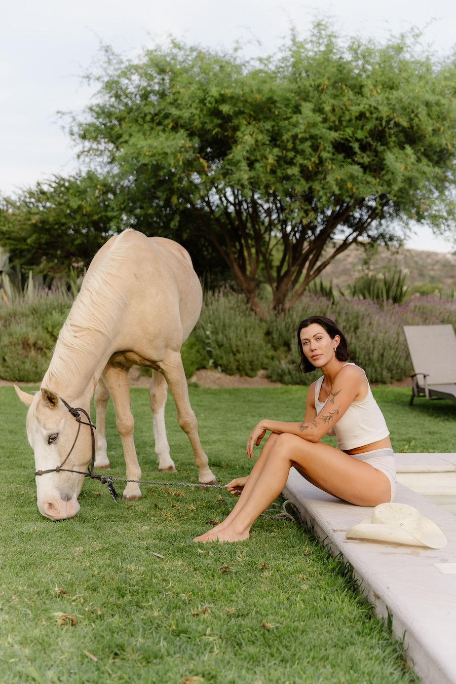 Barefoot bride sits beside horse grazing on lush green grass for a timeless photo on her wedding day