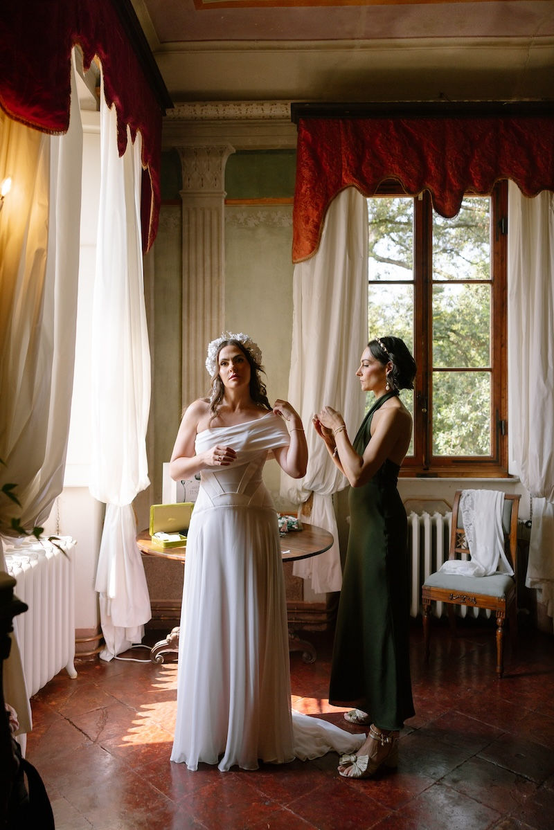 Bride adjusting white dress with bridesmaid in sunlit room.
