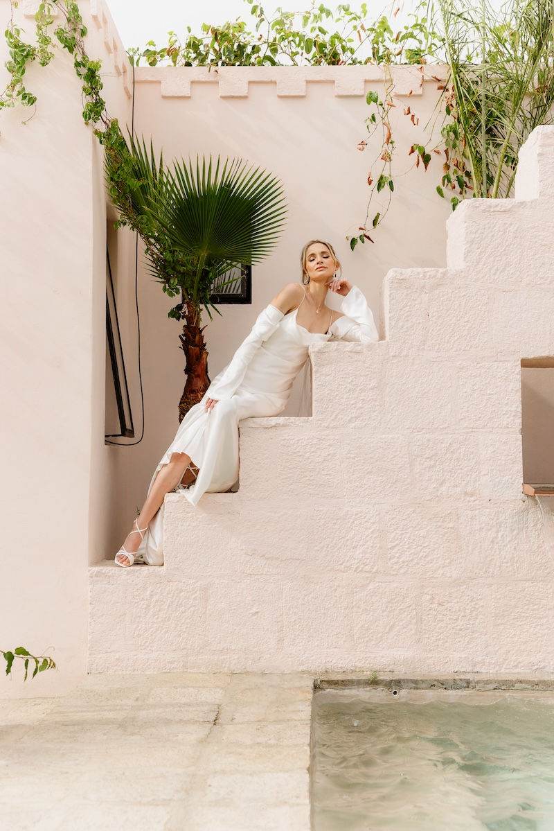 A young bride in white dress relaxes on light pink stairs by pool, posing for a stylish photo for Jo and Glo