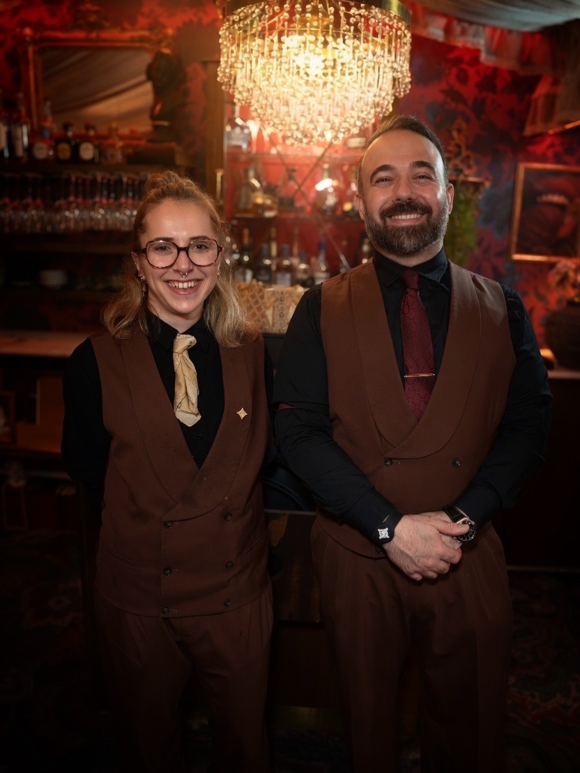 Smilende bartendere i brune vester foran en lyskrone.