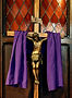 Wooden crucifix with antique corpus, draped with purple fabric in front of a wooden panel with orange and red diamond patterned stainglass inset on the top