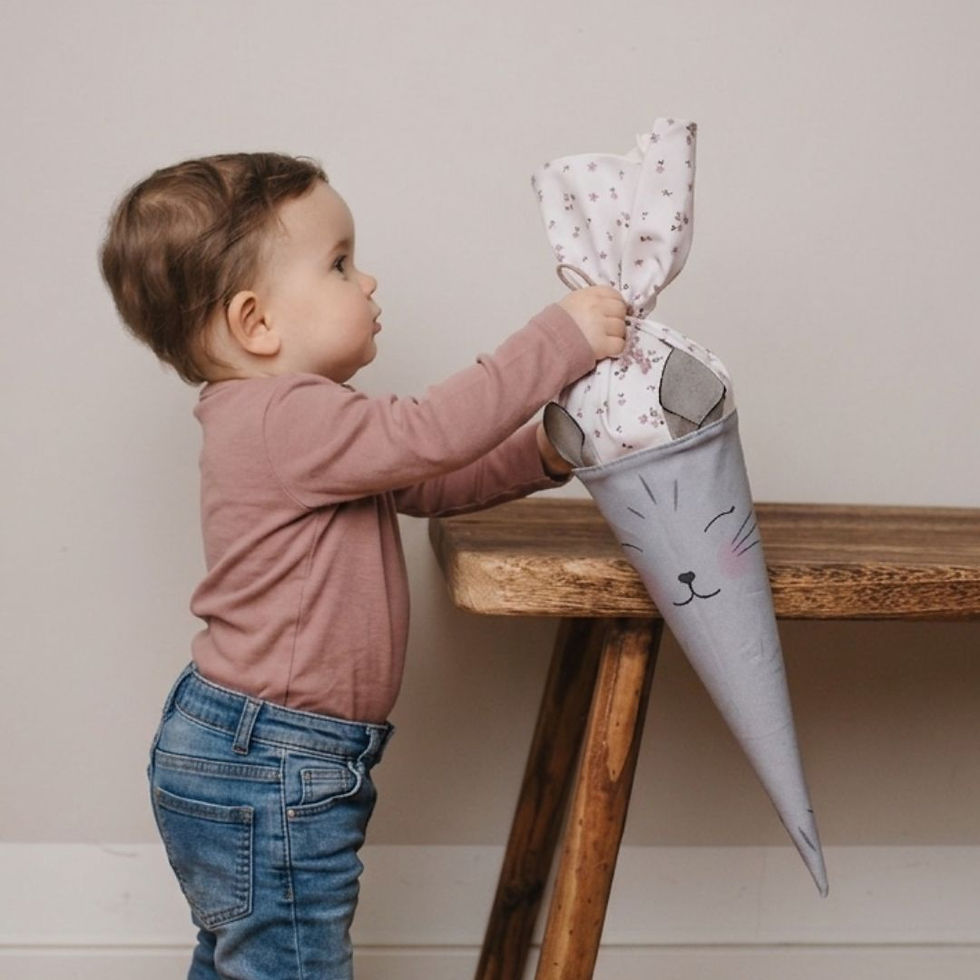 Miniaturbild: Kleinkind greift nach einer Geschwistertüte aus Stoff mit Katzenmotiv auf einer Holzbank