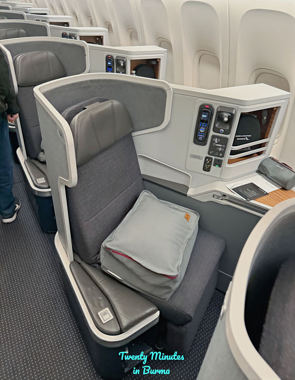 Gray airline business class cabin with plush seats and pillows, control panels, and privacy dividers. White and gray tones dominate the setting. American Airlines flight from LAX to Heathrow