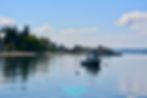 Boat anchored on calm water with houses and trees lining the distant shore under a blue sky with scattered clouds. West Seattle.