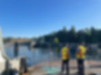 Two people in reflective vests stand on a ferry, facing a dock with trees and clear sky in the background. Visible text: WSF. Ferry arriving in Point Defiance, Tacoma, Washington from Vashon Island.