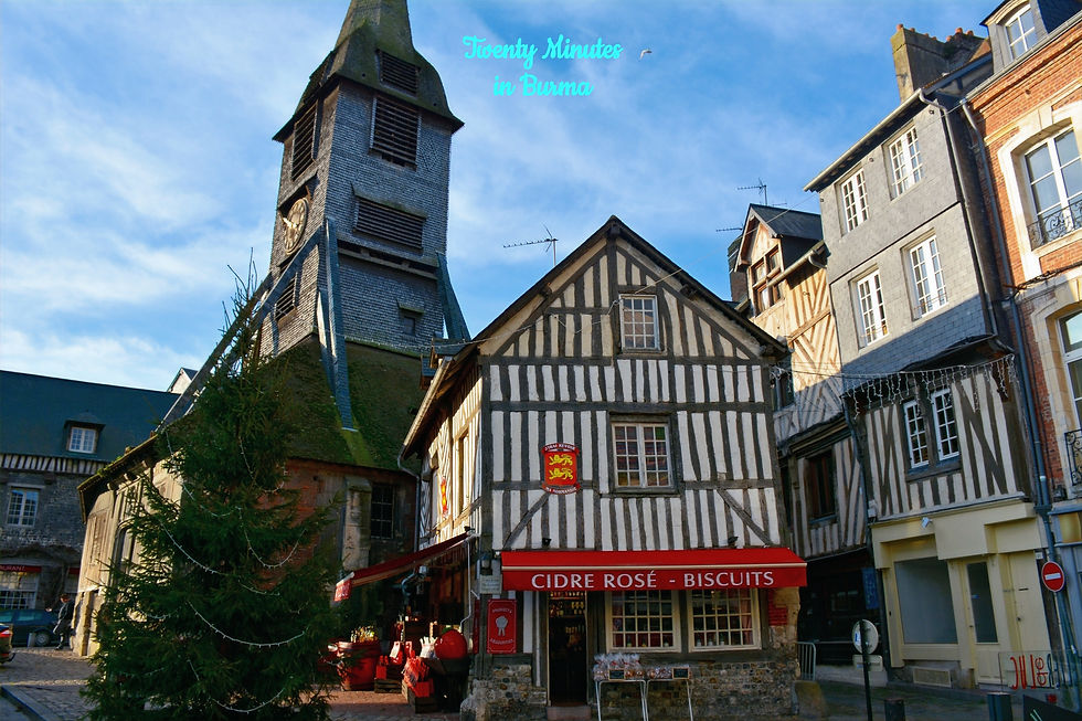 Quaint village scene with a timber-framed house, church tower, and a decorated Christmas tree. Sign reads "Cidre Rosé - Biscuits." Honfleur, France