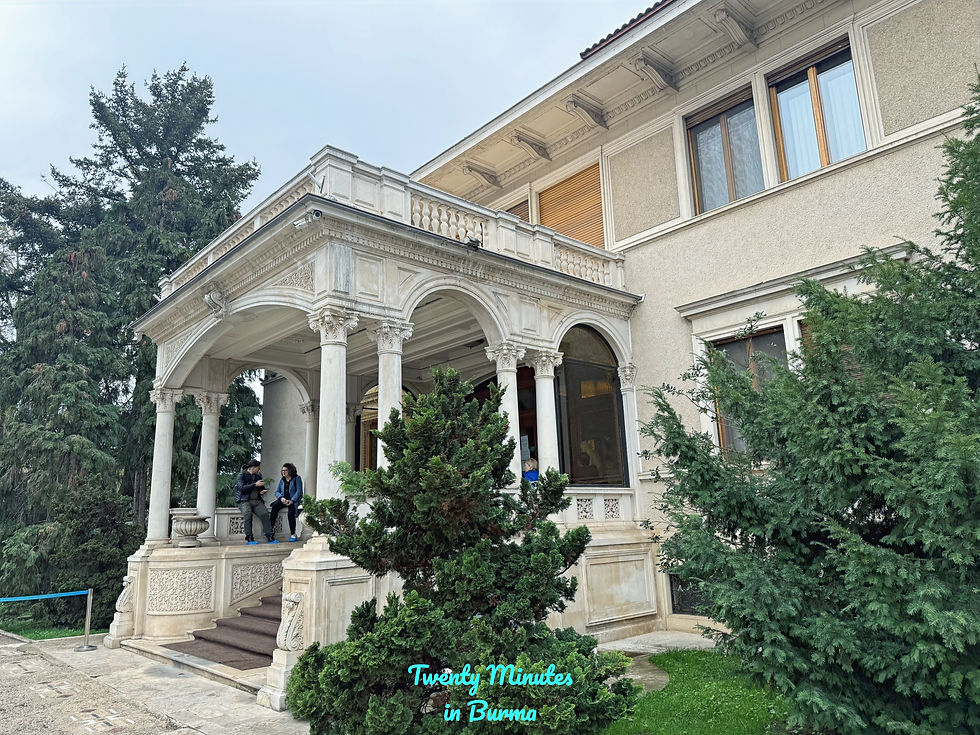 A classic building with ornate columns and an archway. Two people sit on steps, surrounded by greenery. Overcast sky. Casa Ceausescu, Bucharest, Romania