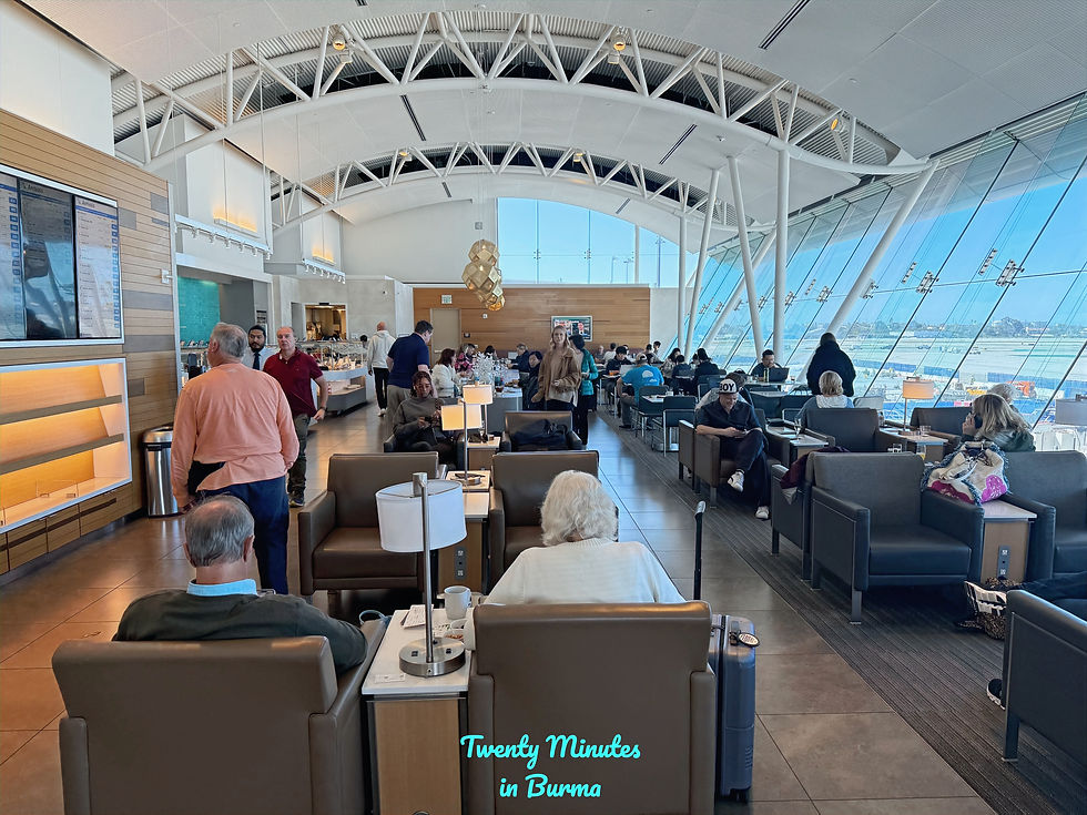 People in an airport lounge with large windows showing the runway. Some are seated, others walking. Modern design, bright natural light. American Airlines Flagship Lounge LAX
